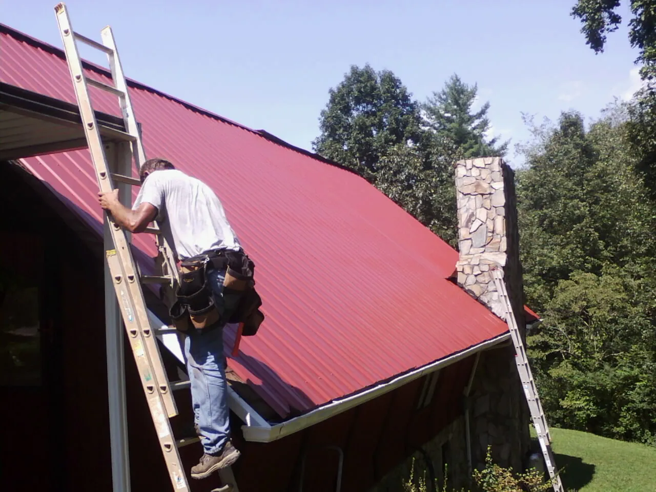 Roofer on a ladder beside a red metal roof and stone chimney — Soffit & Fascia Repair by Roofing Brothers in Moses Lake